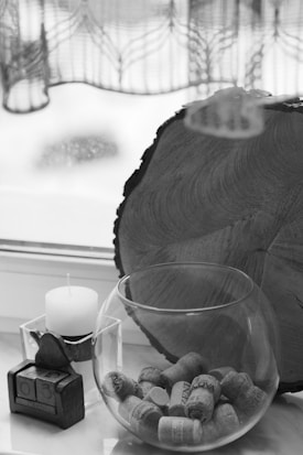 A large, round wood slice stands vertically next to a windowsill. In front of it is a glass bowl filled with wine corks. To the left, there is a white candle sitting in a square holder. Additionally, there is a small calendar display made of wood featuring a heart-shaped ornament.