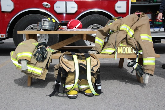 A wooden bench in front of a fire engine holds firefighter gear, including helmets, jackets, gloves, and boots. The items are neatly arranged, indicating readiness. The fire engine in the background is red with silver elements.