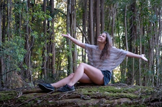 A woman sits on a moss-covered log in a forest with her arms outstretched, seemingly embracing nature. Tall trees with thick trunks and abundant green foliage surround her.
