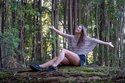 A woman sits on a moss-covered log in a forest with her arms outstretched, seemingly embracing nature. Tall trees with thick trunks and abundant green foliage surround her.