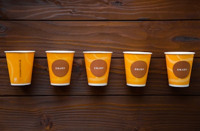 Five orange paper cups are arranged in a neat row on a wooden surface. Each cup features a brown circular design with the word 'ENJOY' in white letters. The wood has a rich, dark grain that contrasts with the bright color of the cups.