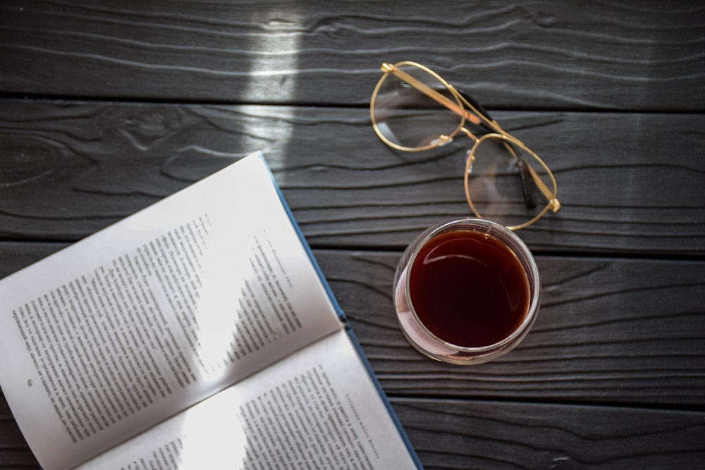 book and eyeglasses beside beverage in glass on gray wood planks