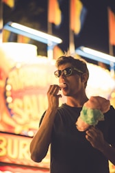 A person wearing sunglasses enjoys cotton candy at a brightly lit carnival. The background features colorful lights and blurred signage, creating a lively atmosphere.