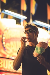 A person wearing sunglasses enjoys cotton candy at a brightly lit carnival. The background features colorful lights and blurred signage, creating a lively atmosphere.
