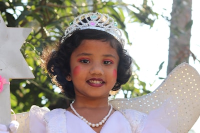 A young child wearing a sparkling silver tiara and pink makeup, dressed in a white costume with a pearl necklace, appears to be portraying a fairy or princess. The background consists of green foliage with sunlight filtering through.
