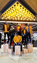 Mannequins dressed in coordinated fashion outfits, predominantly in shades of yellow and black, are displayed on wooden platforms in a well-lit store. The ceiling features an array of hanging light bulbs that add to the contemporary ambiance.