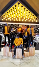 Mannequins dressed in coordinated fashion outfits, predominantly in shades of yellow and black, are displayed on wooden platforms in a well-lit store. The ceiling features an array of hanging light bulbs that add to the contemporary ambiance.