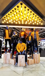 Mannequins dressed in coordinated fashion outfits, predominantly in shades of yellow and black, are displayed on wooden platforms in a well-lit store. The ceiling features an array of hanging light bulbs that add to the contemporary ambiance.