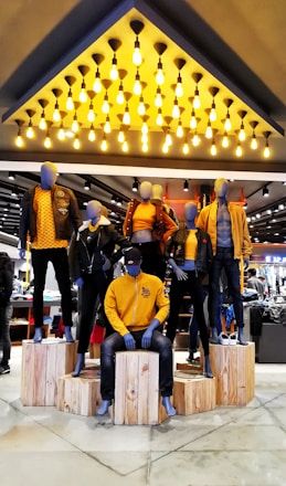 Mannequins dressed in coordinated fashion outfits, predominantly in shades of yellow and black, are displayed on wooden platforms in a well-lit store. The ceiling features an array of hanging light bulbs that add to the contemporary ambiance.