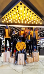 Mannequins dressed in coordinated fashion outfits, predominantly in shades of yellow and black, are displayed on wooden platforms in a well-lit store. The ceiling features an array of hanging light bulbs that add to the contemporary ambiance.