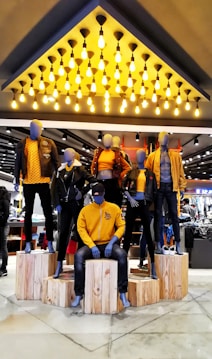Mannequins dressed in coordinated fashion outfits, predominantly in shades of yellow and black, are displayed on wooden platforms in a well-lit store. The ceiling features an array of hanging light bulbs that add to the contemporary ambiance.