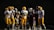 A group of American football players from two different teams, wearing distinct uniforms of yellow and maroon, gather together on a field. The scene is set at night with the players' helmets and gear clearly visible, suggesting a game or practice session. Trees are visible in the background under the night sky.
