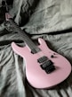A pink electric guitar is placed on a greyish textured fabric background. The guitar features a sleek design with a black bridge and pickups.