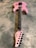 A pink electric guitar lies on a textured gray surface. It features a sleek body design with black hardware, including knobs and pickups. The neck and fretboard are dark-colored, and the headstock is marked with a signature or logo in black.