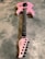 A pink electric guitar lies on a textured gray surface. It features a sleek body design with black hardware, including knobs and pickups. The neck and fretboard are dark-colored, and the headstock is marked with a signature or logo in black.