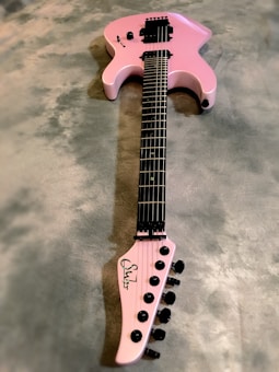 A pink electric guitar lies on a textured gray surface. It features a sleek body design with black hardware, including knobs and pickups. The neck and fretboard are dark-colored, and the headstock is marked with a signature or logo in black.