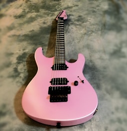 A pink electric guitar with a sleek body design is positioned on a textured gray surface. The guitar features black hardware, including pickups and tuning pegs, and has a contemporary look with its smooth, glossy finish.