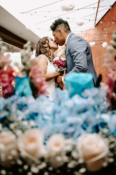 A couple is kissing under a decorated ceiling, surrounded by vibrant floral arrangements. The man is wearing a blue suit and the woman is in a white dress, holding a bouquet of pink flowers. The setting has a warm, intimate atmosphere with light strings hanging above.