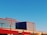 A modern building with a geometric design featuring large metal panels and a striking color scheme of red and blue. The structure has a flat roof and expansive windows, positioned against a clear blue sky. The sunlight casts subtle shadows on the building's surface, adding depth and contrast.
