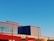 A modern building with a geometric design featuring large metal panels and a striking color scheme of red and blue. The structure has a flat roof and expansive windows, positioned against a clear blue sky. The sunlight casts subtle shadows on the building's surface, adding depth and contrast.