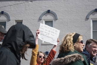 A group of people stand outdoors, participating in a march or protest. One person prominently holds up a sign with messages demanding action against school shootings. They are dressed in casual winter clothing, including jackets and sunglasses. The background features a white brick building with windows.
