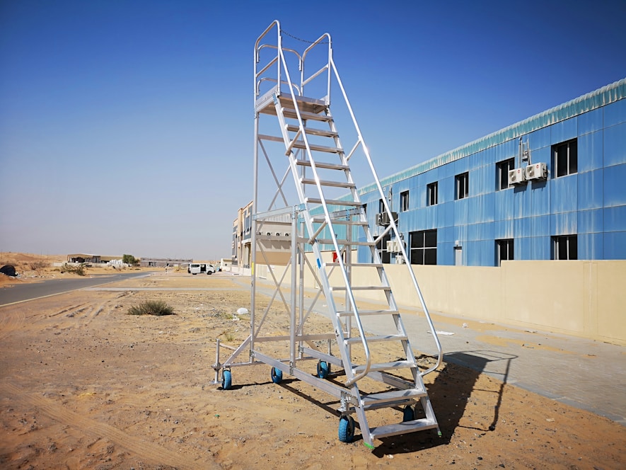 A portable metal staircase with blue wheels stands on sandy terrain next to a building with blue-tinted windows and multiple air conditioning units. The background features a road leading towards other buildings and a vast desert landscape under a clear blue sky.