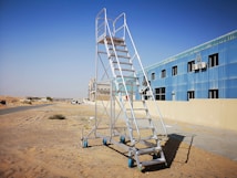 A portable metal staircase with blue wheels stands on sandy terrain next to a building with blue-tinted windows and multiple air conditioning units. The background features a road leading towards other buildings and a vast desert landscape under a clear blue sky.