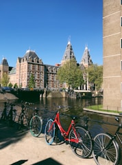 A scenic view features several bicycles lined up along a canal with historic, ornate brick buildings in the background. The clear blue sky and the lush green trees add vibrancy to the scene, enhanced by the sunlight.