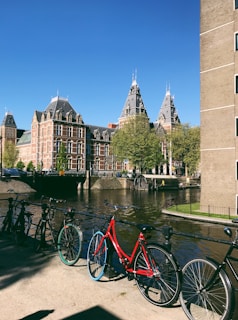 A scenic view features several bicycles lined up along a canal with historic, ornate brick buildings in the background. The clear blue sky and the lush green trees add vibrancy to the scene, enhanced by the sunlight.