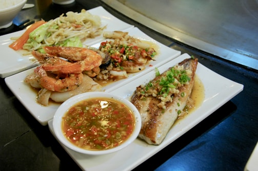 A variety of seafood dishes are presented on white plates, including grilled shrimp, fish with sauce and garnishing, and stir-fried vegetables. A small bowl of dipping sauce with chopped herbs and chili is also visible.