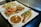 A variety of seafood dishes are presented on white plates, including grilled shrimp, fish with sauce and garnishing, and stir-fried vegetables. A small bowl of dipping sauce with chopped herbs and chili is also visible.