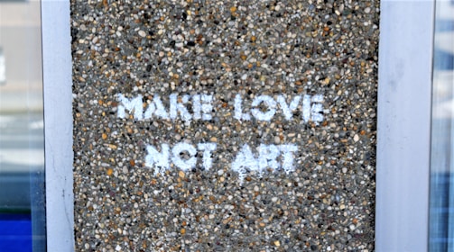 A textured concrete wall with the phrase 'MAKE LOVE NOT ART' stenciled in white letters. The surrounding area is framed by metal or glass structures.