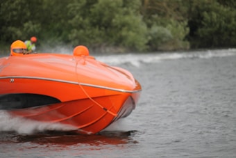 An orange speedboat with two people wearing bright orange helmets moves swiftly across a body of water. In the background, lush greenery lines the shore, and the water is slightly choppy due to the boat's speed.