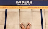A pair of feet wearing flip flops stands on a platform with bilingual safety warnings and a marked yellow line indicating where to wait for a local train. Blue lines and tiles form the background, enhancing platform safety instructions.