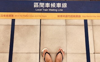 A pair of feet wearing flip flops stands on a platform with bilingual safety warnings and a marked yellow line indicating where to wait for a local train. Blue lines and tiles form the background, enhancing platform safety instructions.
