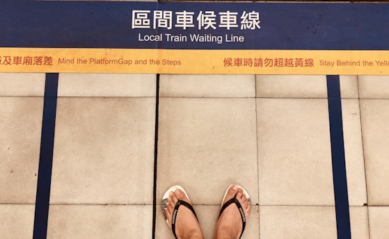 A pair of feet wearing flip flops stands on a platform with bilingual safety warnings and a marked yellow line indicating where to wait for a local train. Blue lines and tiles form the background, enhancing platform safety instructions.