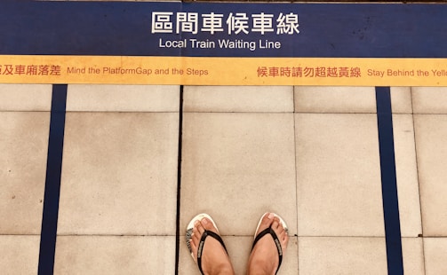 A pair of feet wearing flip flops stands on a platform with bilingual safety warnings and a marked yellow line indicating where to wait for a local train. Blue lines and tiles form the background, enhancing platform safety instructions.