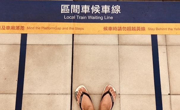 A pair of feet wearing flip flops stands on a platform with bilingual safety warnings and a marked yellow line indicating where to wait for a local train. Blue lines and tiles form the background, enhancing platform safety instructions.
