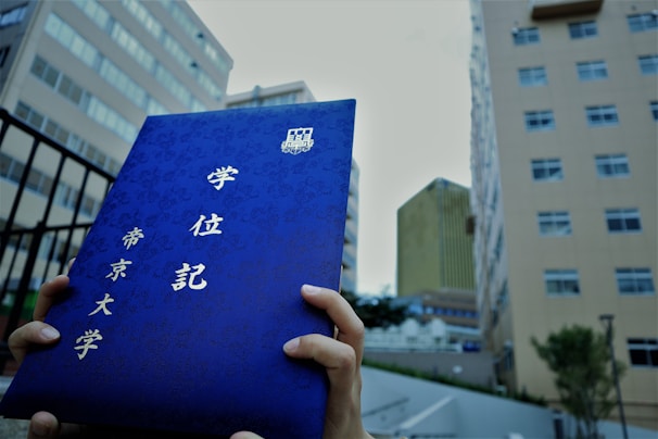 A person holds a blue certificate folder with Japanese characters against the backdrop of tall, urban buildings. The characters are embossed in gold, and the architecture in the background suggests a city environment.