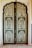 An intricately designed double door features a detailed floral pattern with symmetrical paneling. The use of green and cream colors highlights the botanical motifs, enclosed within an ornate border. The arched top adds an architectural elegance to the overall design.