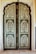 An intricately designed double door features a detailed floral pattern with symmetrical paneling. The use of green and cream colors highlights the botanical motifs, enclosed within an ornate border. The arched top adds an architectural elegance to the overall design.