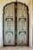 An intricately designed double door features a detailed floral pattern with symmetrical paneling. The use of green and cream colors highlights the botanical motifs, enclosed within an ornate border. The arched top adds an architectural elegance to the overall design.