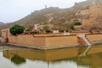 A historical fort situated on a hillside with large sandstone walls reflecting in a body of water. The landscape includes arid terrain with sparse vegetation and a few trees. The architecture features classic fort design with parapets and battlements.