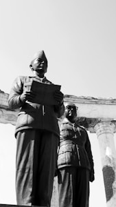 Two statues of men in military uniforms stand prominently with one holding a document. The figures are set against a clear sky, and there is a partial view of a stone or concrete structure with columns in the background.