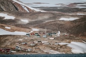 A small, remote settlement nestled among rocky, snow-dotted hills. The area features a collection of colorful buildings, including red, green, and yellow structures. There is a large satellite dish and other equipment indicating a research facility or observatory. The landscape is barren, with patches of snow and ice visible on the ground.