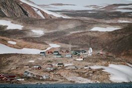 A small, remote settlement nestled among rocky, snow-dotted hills. The area features a collection of colorful buildings, including red, green, and yellow structures. There is a large satellite dish and other equipment indicating a research facility or observatory. The landscape is barren, with patches of snow and ice visible on the ground.