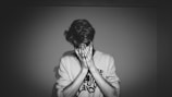 A person stands against a plain background, covering their face with their hands, giving an impression of distress or contemplation. The lighting is dramatic, casting deep shadows, creating a stark contrast in the monochromatic setting.