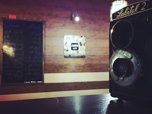 A vintage Lubitel 2 camera is prominently displayed in the foreground on a dark surface. The background features a wooden wall with a chalkboard filled with handwritten script and a square artwork with abstract design that says 'ART' in the center.