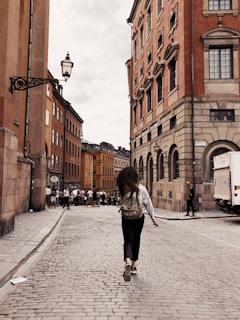 A person with long hair and a backpack walks down a cobblestone street lined with colorful, historic buildings. The scene has an urban, lively feel with several people gathered in the distance. The architecture features ornate details and warm tones, typical of European cityscapes.