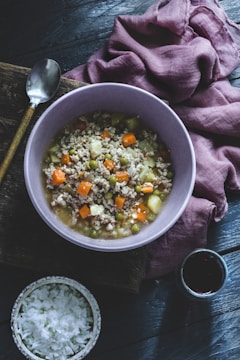 A bowl filled with a hearty soup containing ground meat, carrots, peas, and potatoes is placed on a dark wooden surface. Next to it is a small bowl of rice and a glass of red wine. A spoon with a wooden handle lies nearby on a piece of rustic wood, and a purple cloth is draped casually.